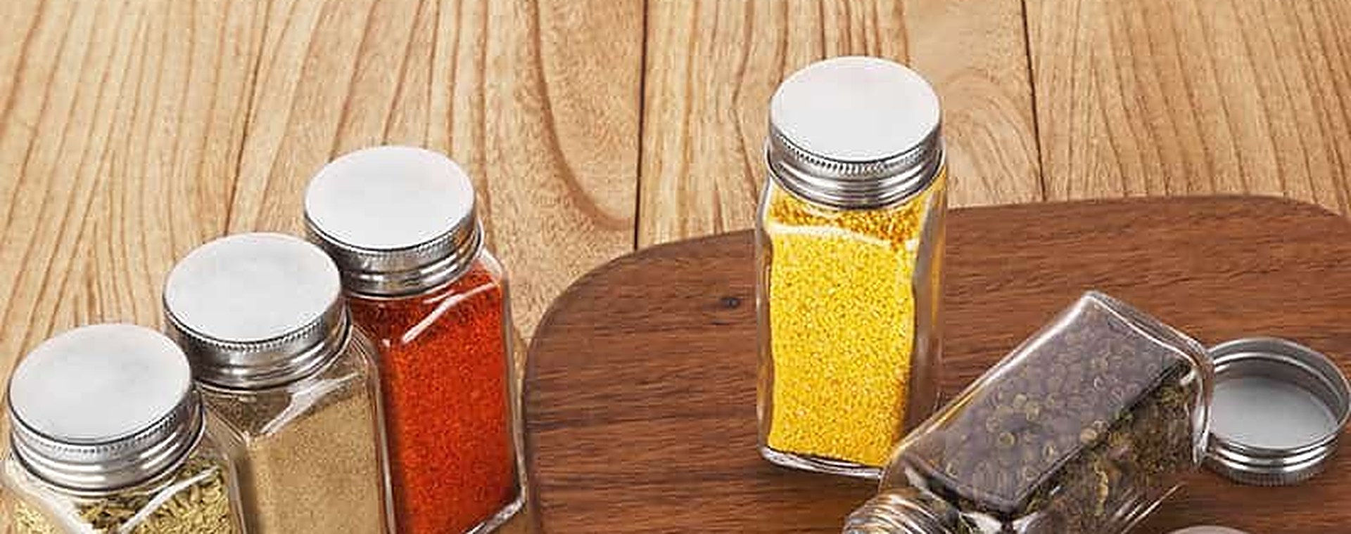 Tarros cuadrados de vidrio para especias con tapa de acero inoxidable sobre una tabla de madera, llenos de pimentón, cúrcuma y pimienta