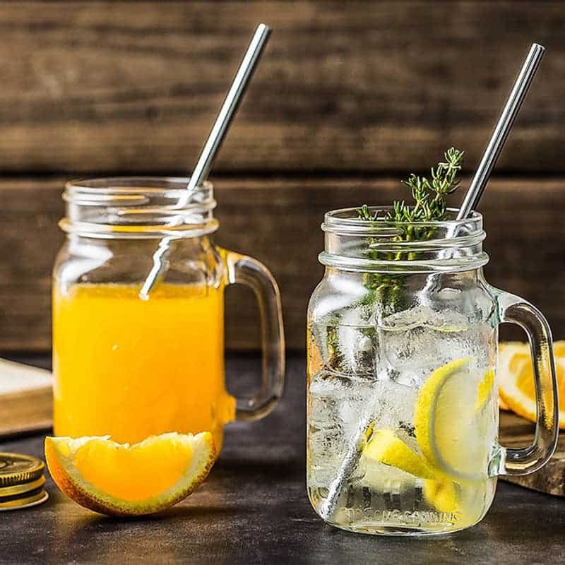 Zwei Mason-Becher mit Henkel und Metall-Trinkhalmen auf einem Holztisch, einer mit Saft, einer mit Zitronenwasser
